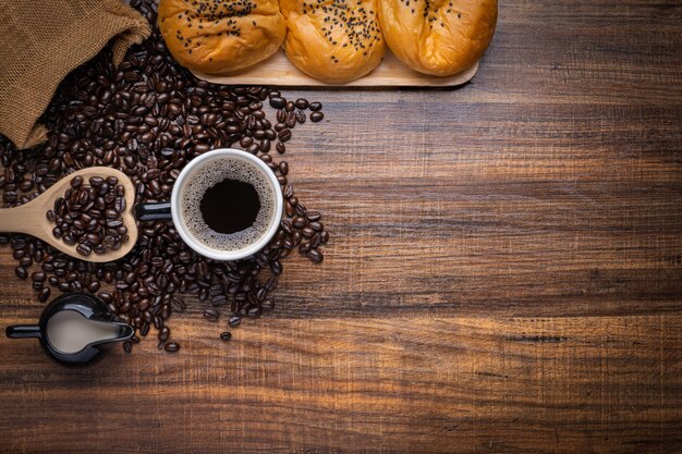 Zwarte en bruine koffiebonen met kop warme drank en brood op houten tafel, bovenaanzicht kopie ruimte