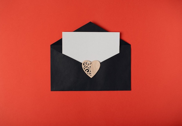 Zwarte envelop met een blanco vel papier erin en een houten hart erop. Valentijnsdag concept. Plat lag, bovenaanzicht.