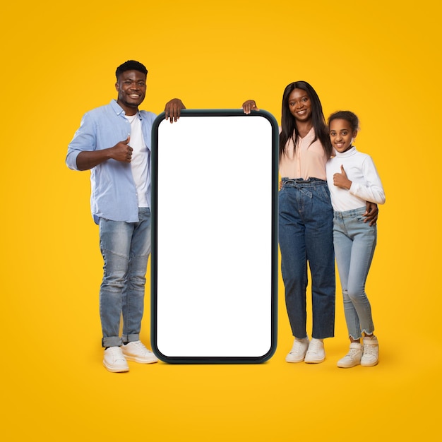 Zwarte familie staande in de buurt van full-length cellpone met leeg scherm gebaren duimen omhoog poseren op gele achtergrond
