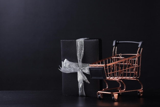 Zwarte geschenkdoos met zilveren lint met mini-trolley geïsoleerd op zwarte achtergrond met kopieerruimte.