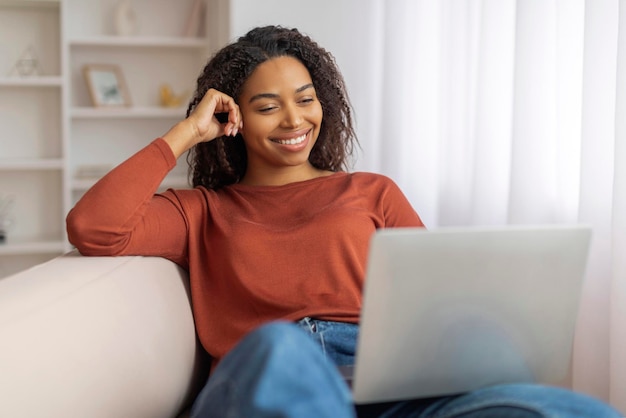 Zwarte glimlachende vrouw zit op de bank met een laptop.