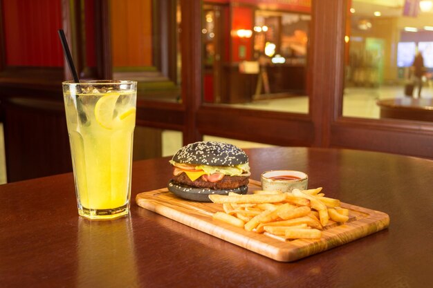 Zwarte hamburgerfrietjes en een kopje tomatensaus op een houten bord met een glas limonade, een schijfje citroen en een buisje