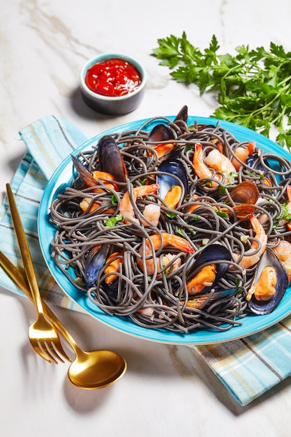 Zwarte inktvisinktpasta met zeevruchten: mosselen en garnalen met witte wijn en knoflooksaus op een blauw bord met gouden bestek op een witmarmeren achtergrond met ketchup en peterselie, bovenaanzicht, close-up