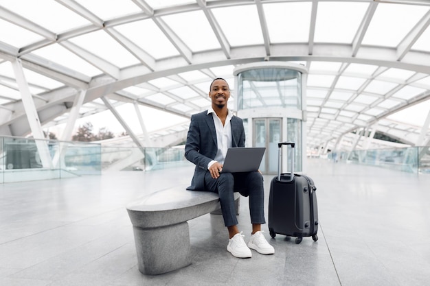 Zwarte jonge zakenman met laptopcomputer zittend op een bankje op de luchthaven