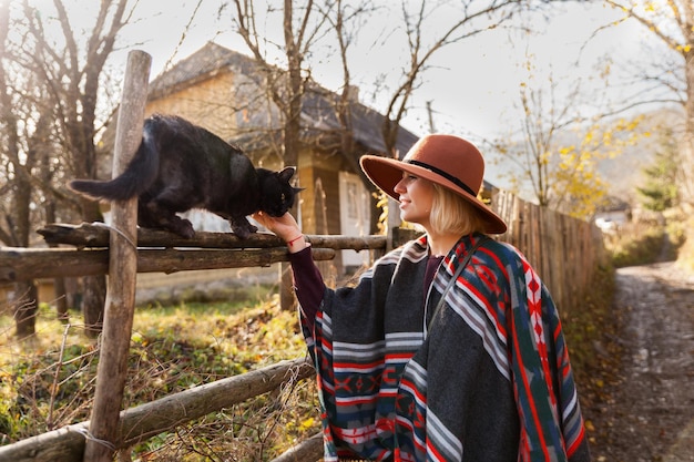 Zwarte kat en stijlvolle reizende vrouw met authentieke poncho in boho-chique stijl en hoed in de buurt van oude houten plattelandsgebouwen Vrouw heeft plezier in de herfsttijdReis- en reislustconcept