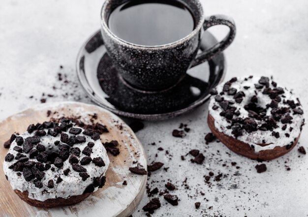 Zwarte koffiekopje met schotel en donuts met zwarte koekjes op stenen keukentafel.