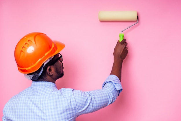 Zwarte man Afro-Amerikaanse verfroller in de hand verft de muur in roze kleur .happy Afrikaanse bouwer schilderij in het huis, zakenman draagt een helm helm. jonge man is aan het schilderen