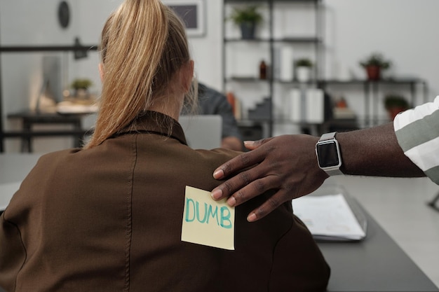 Zwarte man die sticker met het woord dom op de achterkant van een blonde vrouwelijke collega zet