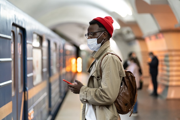 Zwarte man draagt een gezichtsmasker als bescherming tegen het covid-19-virus, terwijl hij in de metro staat en zijn mobiel gebruikt.