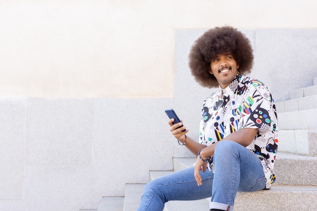 Zwarte man met afro-haar glimlachend zittend op een stenen trap met zijn smartphone in zijn hand naar de zijkant kijkend