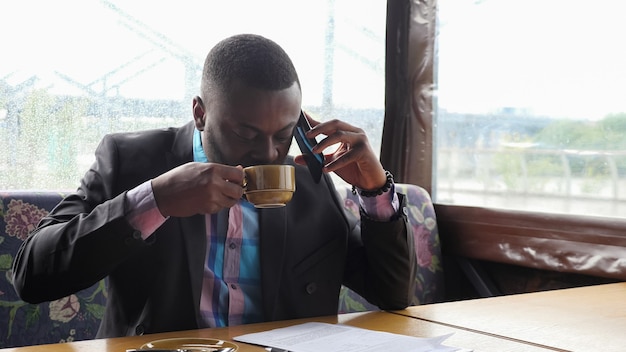 Zwarte man praat op smartphone. Afro-Amerikaanse zakenman roept een mobiele telefoon op die in het zomertentcafé zit. Hij drinkt een kopje koffie aan tafel. Hij draagt hemd en colbert.