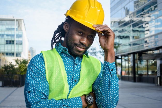 Zwarte mannelijke ingenieur staat buiten en groet met zijn hardhat in de ene hand.