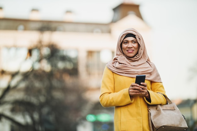 Zwarte moslimvrouw die een hijab draagt en fooi geeft op haar smartphone terwijl ze door het centrum loopt.