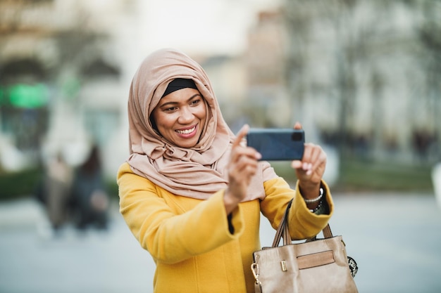 Zwarte moslimvrouw die een hijab draagt en foto's maakt met haar smartphone terwijl ze door het centrum loopt.