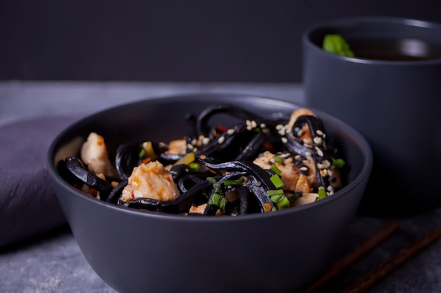 Zwarte pijlinktvisinkt udon noedels met kippenvlees op de zwarte kom en kop thee op donkere steenachtergrond.