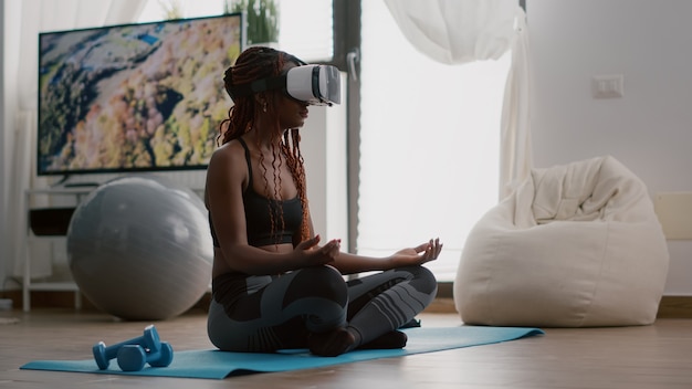 Zwarte trainervrouw die een virtual reality-headset draagt terwijl ze op de yogakaart zit