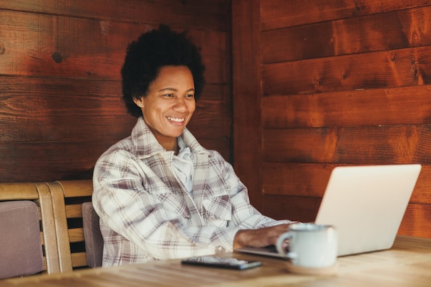 Zwarte vrouw die op laptop werkt terwijl ze vakantietijd doorbrengt in een gezellig houten huis.