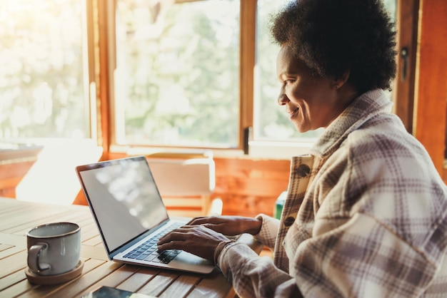 Zwarte vrouw die 's ochtends op een laptop werkt terwijl ze vakantietijd doorbrengt in een gezellig houten huis in een bos.