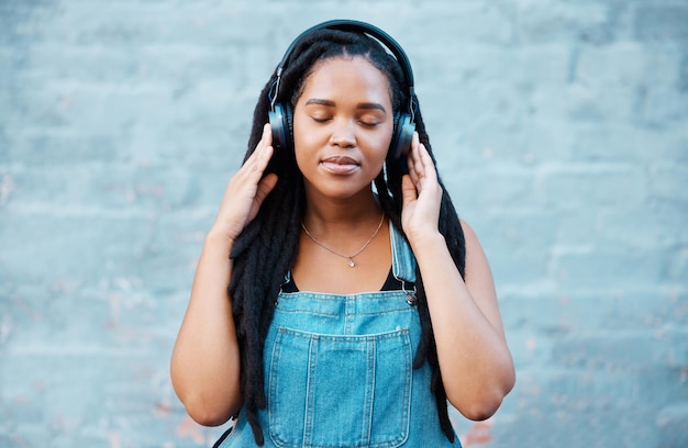 Zwarte vrouw koptelefoon en muziek met een meisje dat zich kalm voelt, ontspannen en tevreden luisterend naar een podcast die buiten tegen de stads- of stadsmuur staat Afrikaanse vrouw ontspannen tijdens bewuste audiostreaming