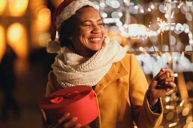 Zwarte vrouw van middelbare leeftijd met sterretjes en kerstcadeaudoos op feestelijke winternacht in de stad.