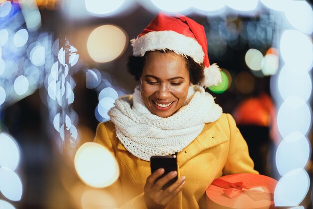 Zwarte vrouw van middelbare leeftijd typen op haar smartphone op de kerstmarkt op feestelijke nacht in de stad.