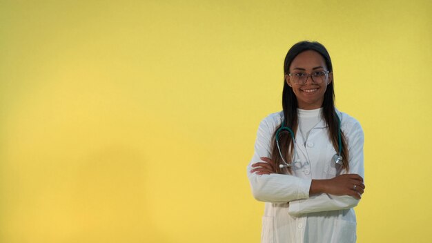 Zwarte vrouwelijke arts die in laboratoriumlaag aan de camera op gele achtergrond glimlacht.