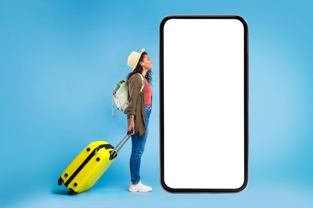 Zwarte vrouwelijke reiziger met koffer en rugzak die naar grote smartphone kijkt met mockup voor online