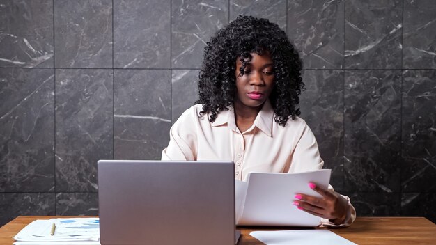 Zwarte zakenvrouw in witte blouse verzamelt papieren kijkt door en typt op laptop zittend aan bruine houten tafel in kantoor met marmeren muur