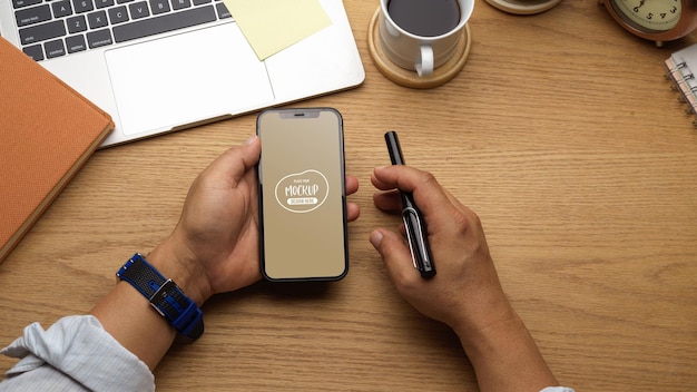 Close up of businessman holding smartphone and pen in his hand on wooden workspace