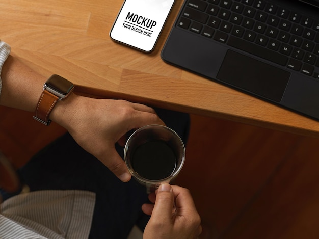 Close up of male hands holding coffee cup at workspace with smartphone mockup