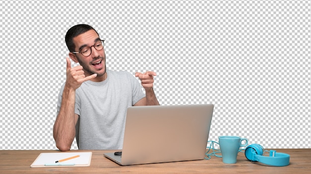 Confident young man sitting at his desk doing a gesture of call with the hand