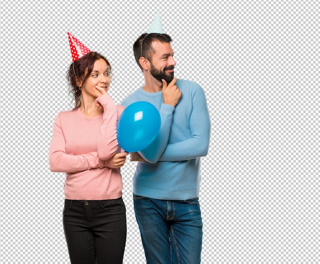 Couple with balloons and birthday hats looking to the side with the hand on the chin