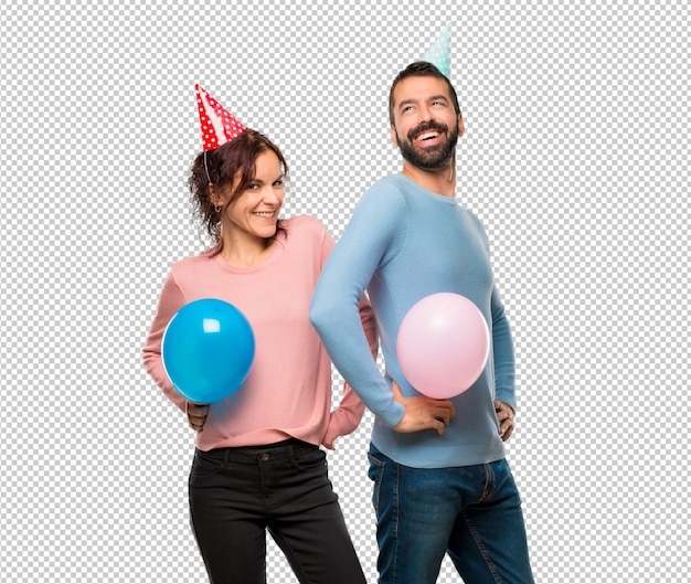 Couple with balloons and birthday hats posing with arms at hip and laughing