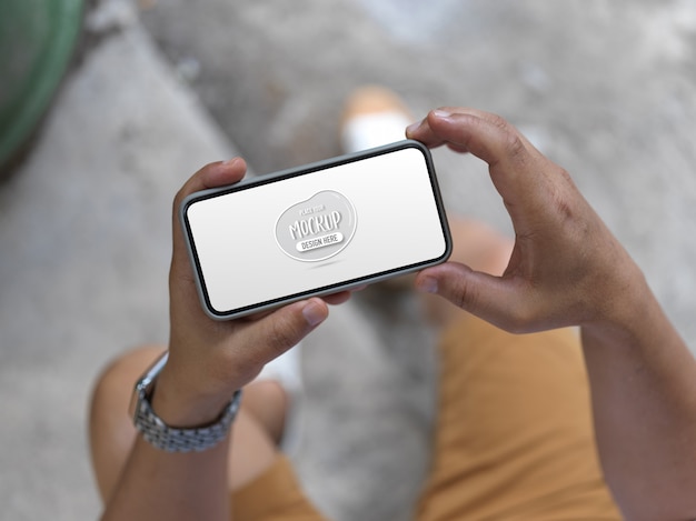 PSD cropped shot of young man holding mock up horizontal smartphone while sitting outdoor