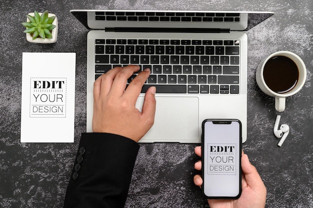 フラットレイトップビュー空白のモックアップスマートフォンディスプレイ若いビジネスマンの手とラップトップコンピューターのイヤホンと植物を備えたオフィスデスクの紙の上の空のスペースあなたのデザインを挿入するための空のスペース