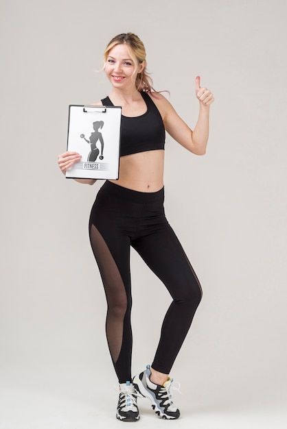 PSD front view of smiley fitness woman holding notepad and giving thumbs up