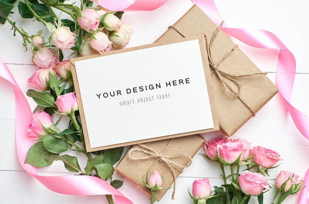 Mockup di biglietto di auguri con scatole regalo e fiori di rose su fondo di legno bianco