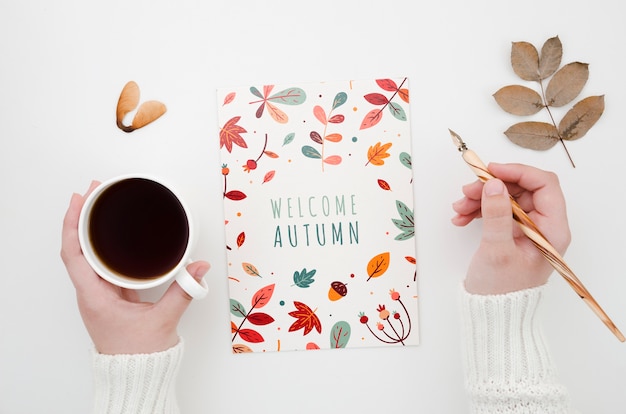 Hands holding coffee and pen next to autumn card 