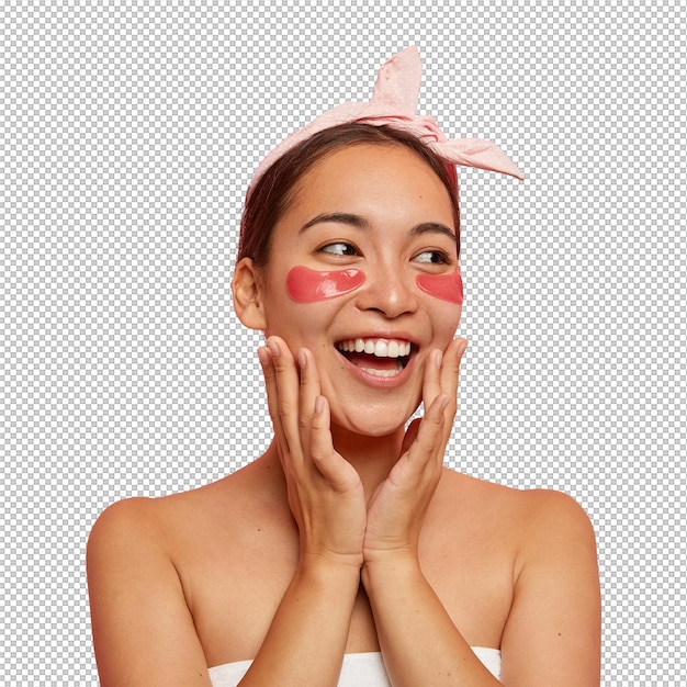 happy young asian woman has eyes cosmetic mask laughs positively touches face gently looks aside conducts beauty treatments in spa salon wears pink headband stands wrapped in towel indoor