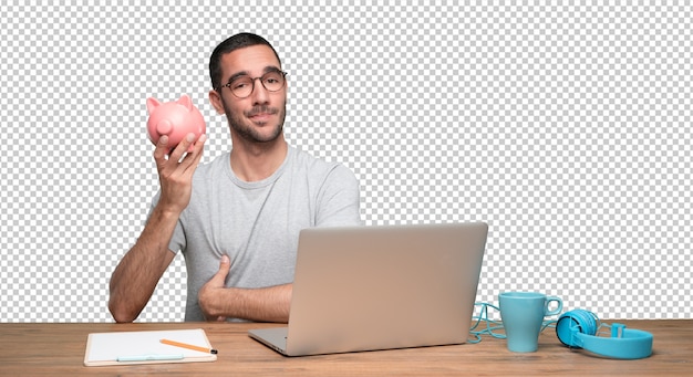 PSD happy young man sitting at his desk and holding a piggy bank