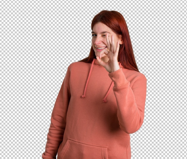 PSD jong roodharigemeisje met roze sweatshirt die een ok teken met vingers tonen terwijl het knipogen van een oog