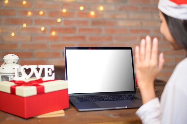 Jonge glimlachende vrouw die rode kerstmanhoed draagt die videogesprek op laptop model maakt