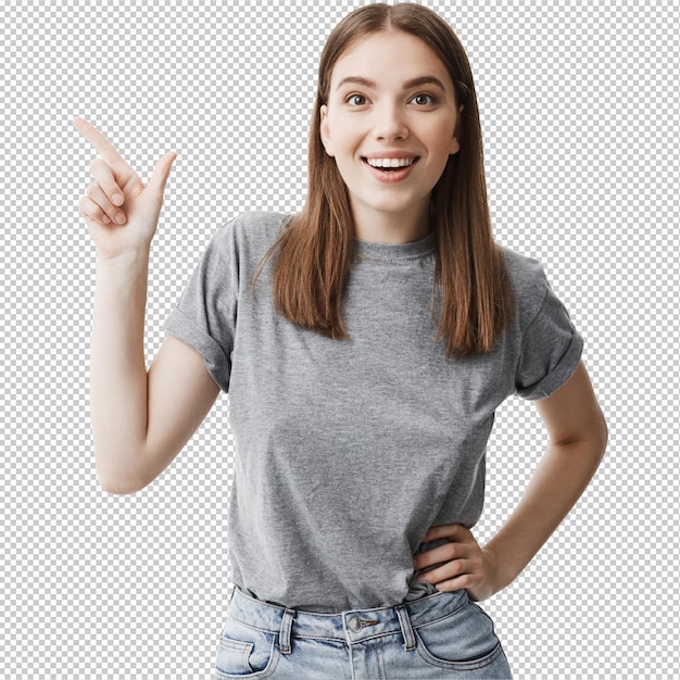 Jonge, scherpe, mooie, donkere jonge vrouw met middelgrote haarlengtes in grijs T-shirt en spijkerbroek die helder glimlacht met een gespannen en gelukkige uitdrukking die naar de witte muur wijst.