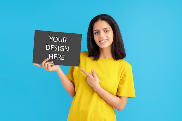 Latin woman with yellow t-shirt holding poster and pointing finger at it, mockup