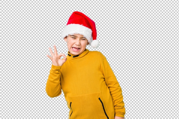 サンタの帽子をかぶってクリスマスの日を祝う少年は目をまばたきし、手でいいジェスチャーを保持します。