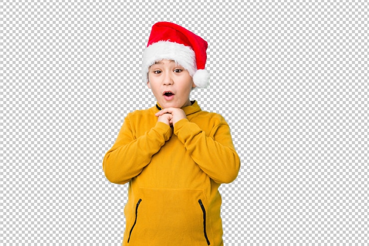 PSD little boy celebrating christmas day wearing a santa hat praying for luck, amazed and opening mouth looking to front.