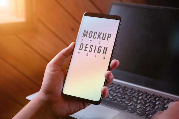 モックアップのスマートフォン画面。ぼやけたノートパソコンで現代のスマートフォンテクノロジーを使用して手を閉じる