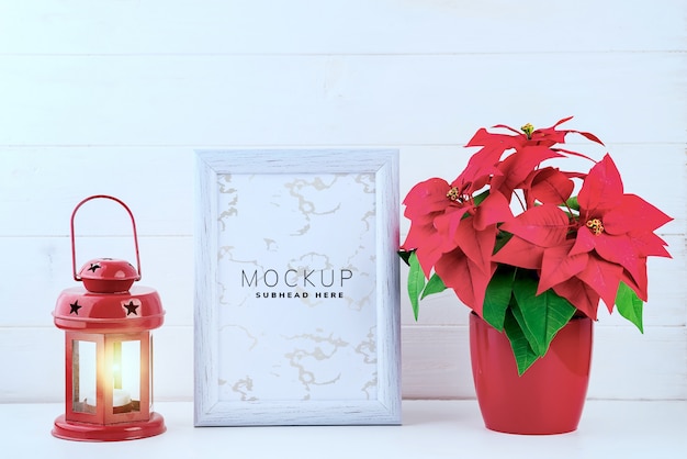 Foto mock up con cornice bianca, stella di natale in un vaso di fiori e lanterna rossa su fondo di legno bianco
