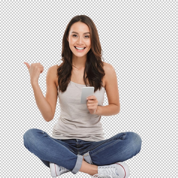 photo of pleased woman with long brown hair sitting with legs crossed on the floor using silver smartphone and pointing away isolated over white wall