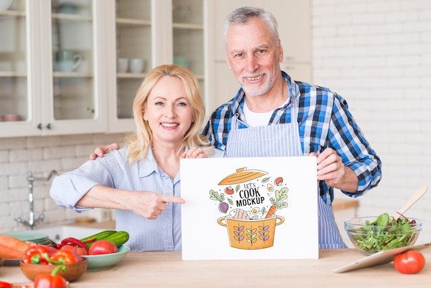 PSD senior couple in kitchen holding paper mock-up